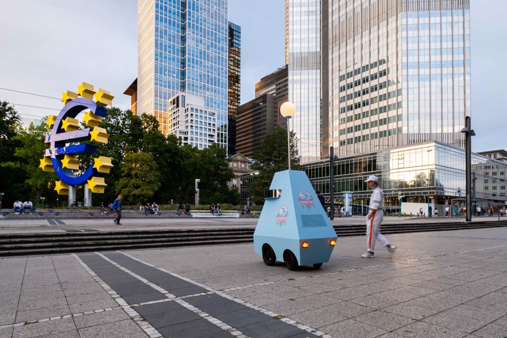 Medium shot of the Willy Brandt Platz in Frankfurt with the Taunusanlage and a big sculpture in the shape of a €-sign being visible in the background, a robot and a person are walking towards the park